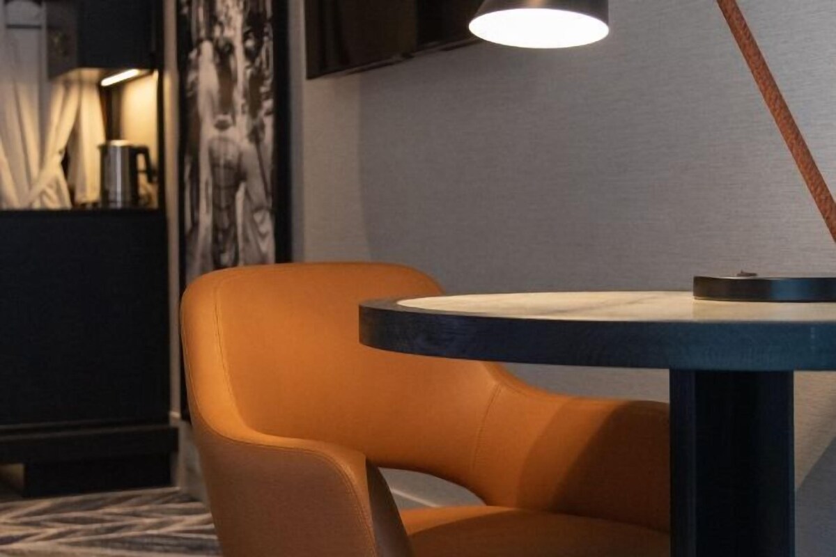 A room with a round table, a single orange chair, a lamp, and a wall-mounted television.