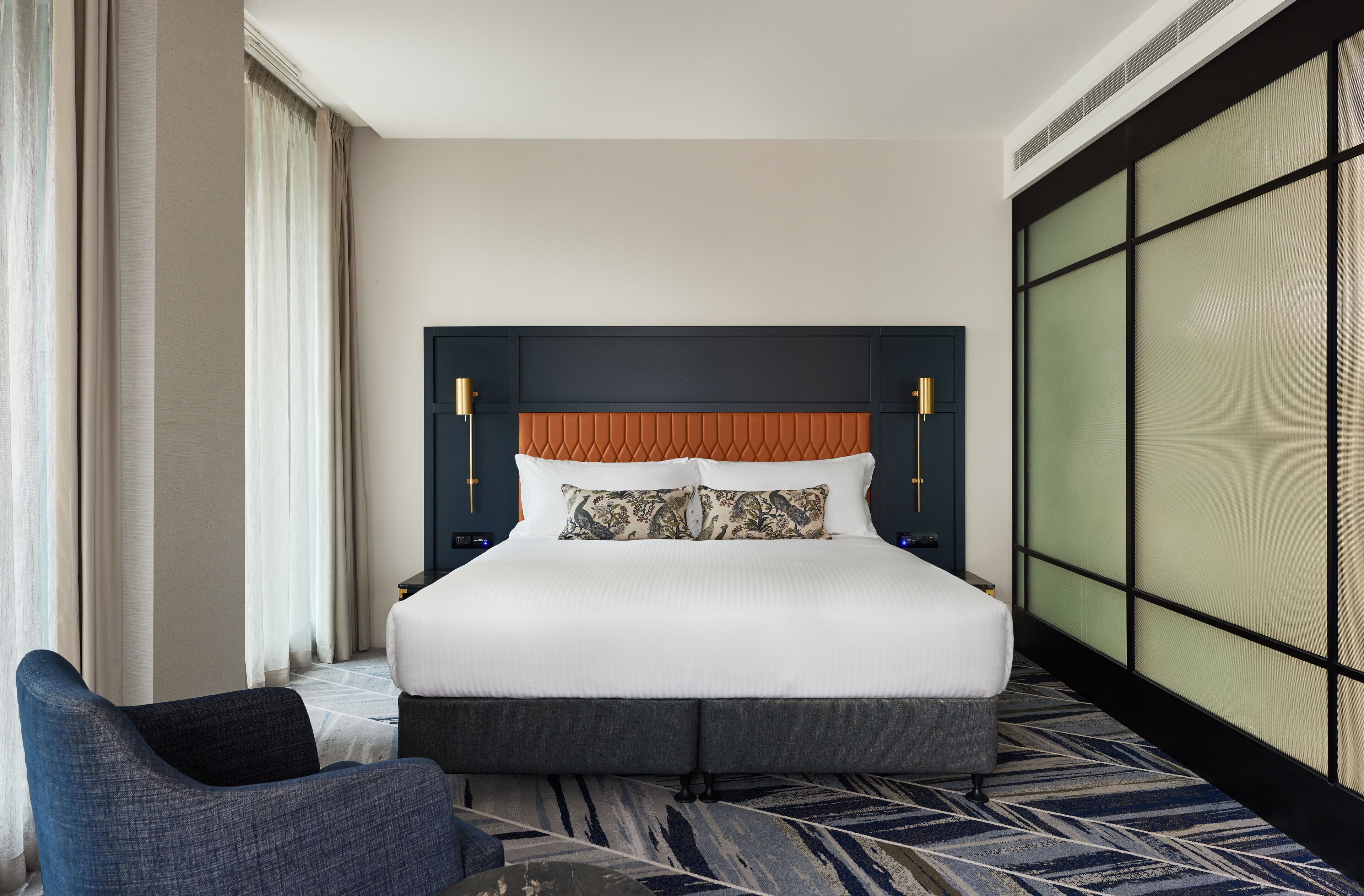 A hotel room with a large bed, a nightstand, a chair, and a view of the outside through the window.