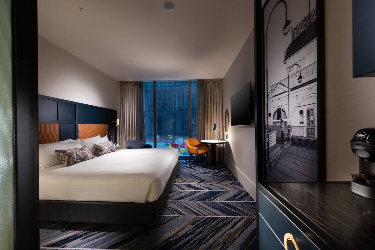 A hotel room with a large bed, a desk, a chair, a window with curtains, and a wall-mounted TV.