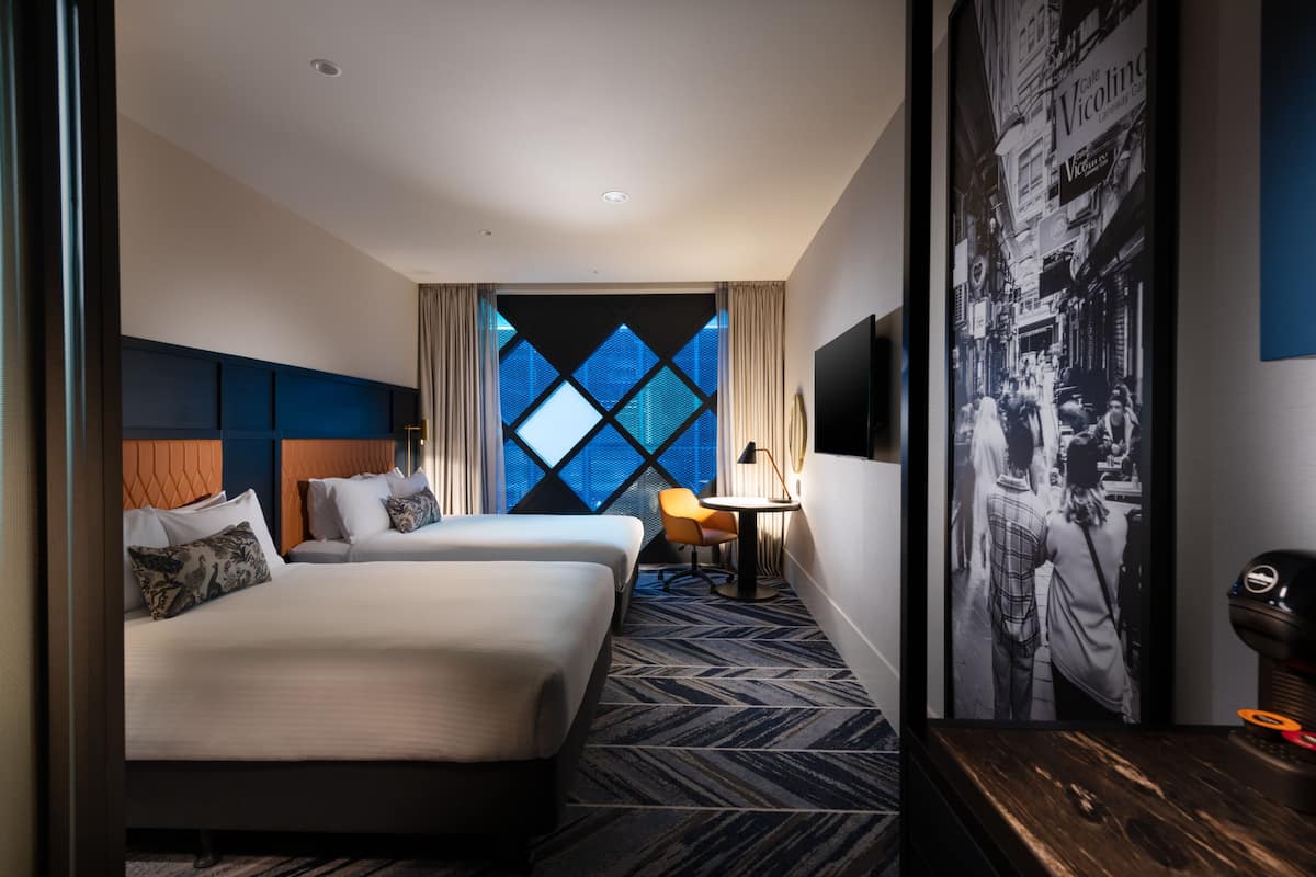 A hotel room with two beds, a window with a geometric pattern, a small table with a lamp, and a wall-mounted TV.