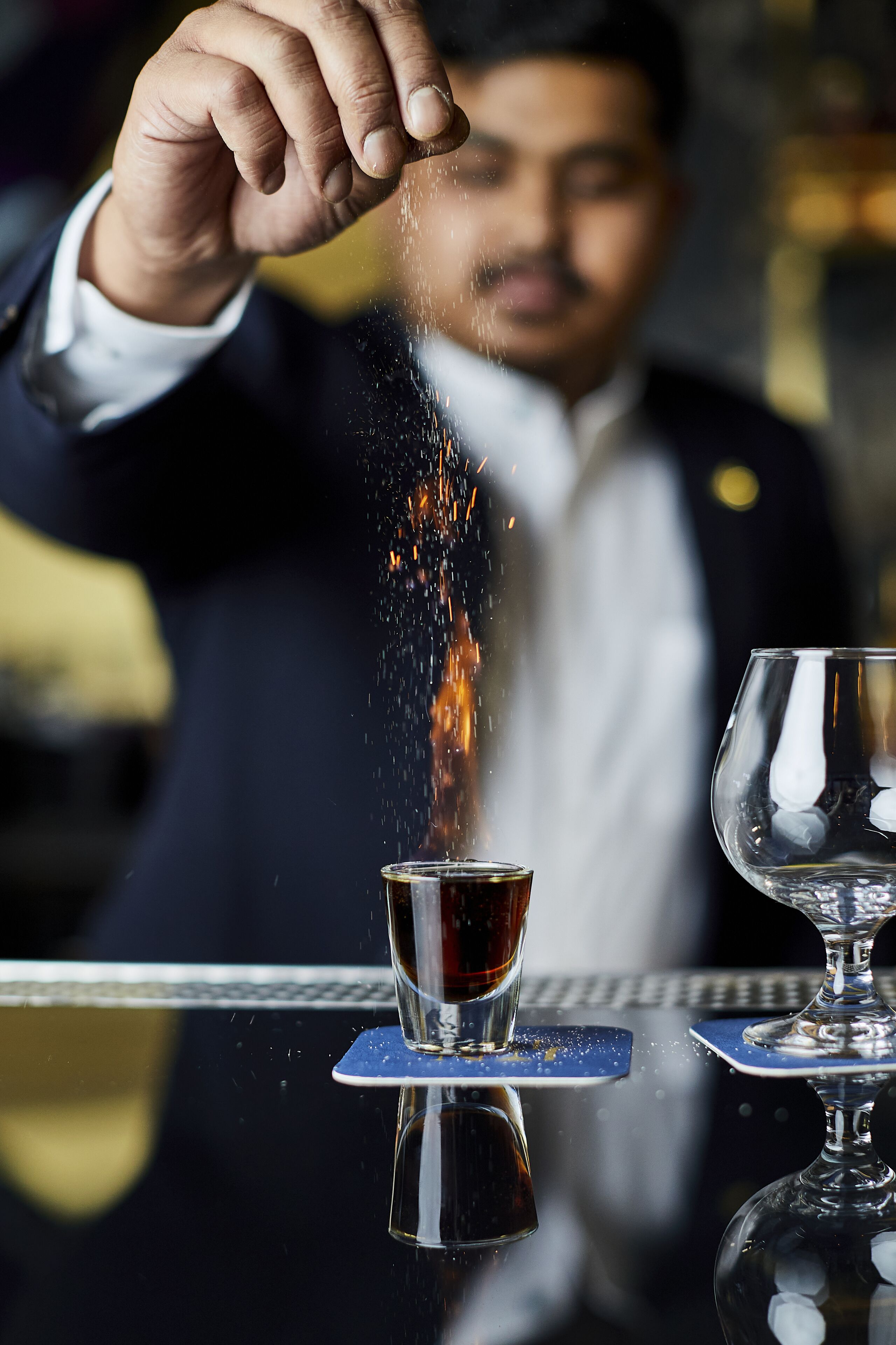 Una persona vertiendo un líquido oscuro en un vaso, con otro vaso de líquido transparente sobre la mesa.