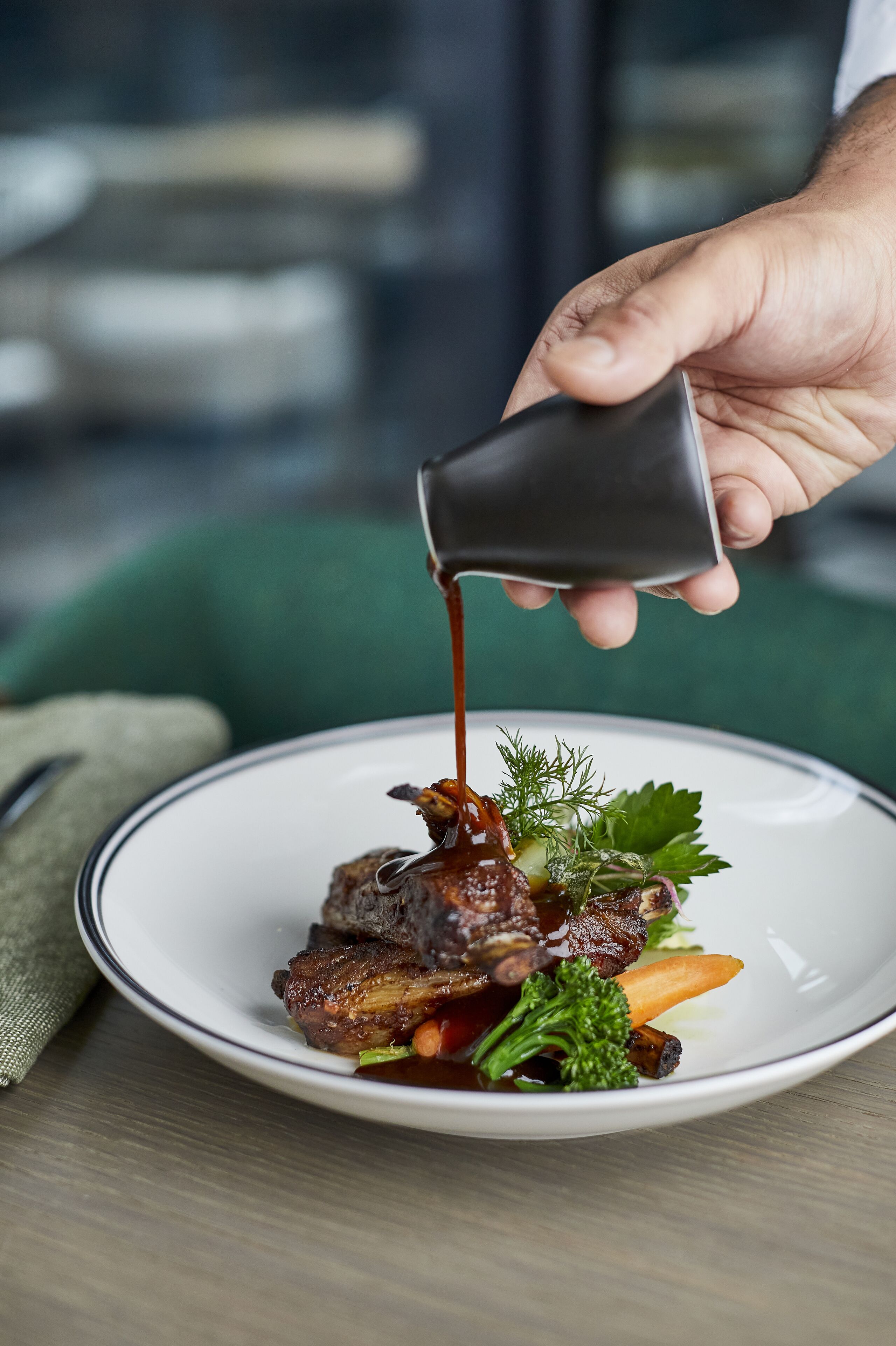 Une main verse de la sauce sur un plat de viande et de légumes, qui se trouve dans une assiette.
