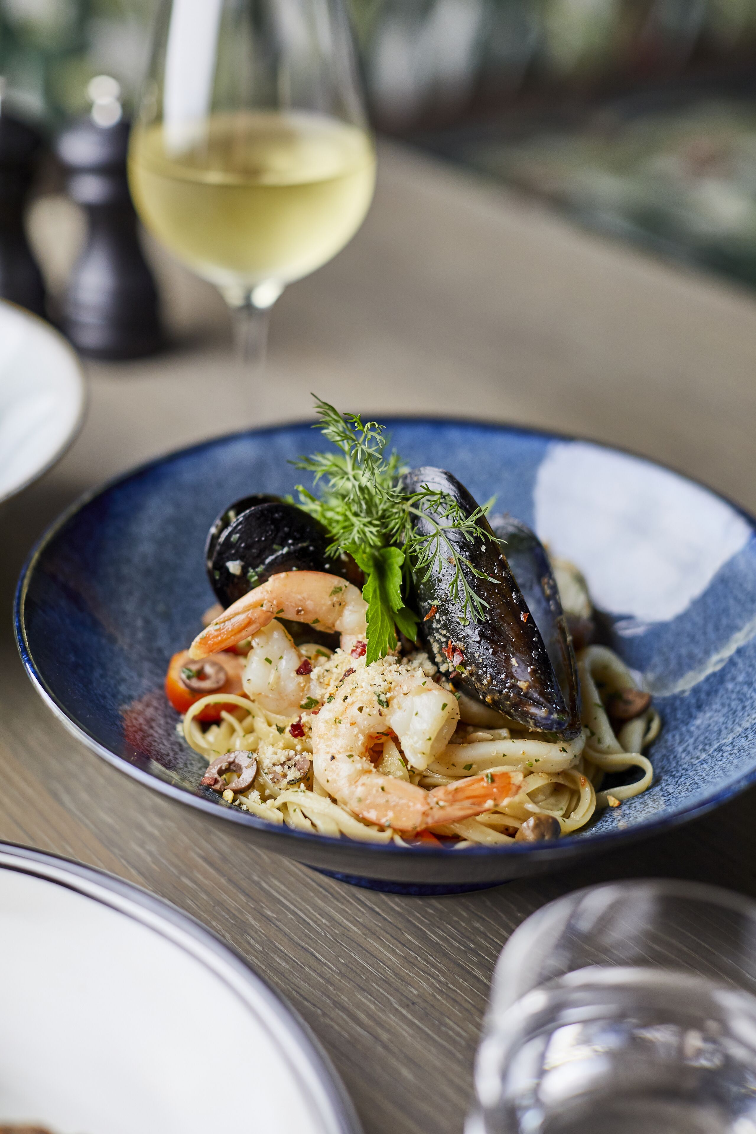 A seafood pasta dish on a blue plate with a glass of white wine in the background.