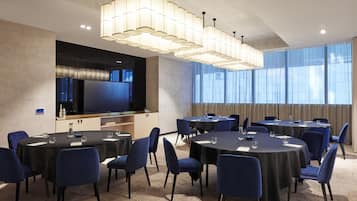 A conference room with multiple round tables, blue chairs, a large flat-screen TV, and a modern chandelier.