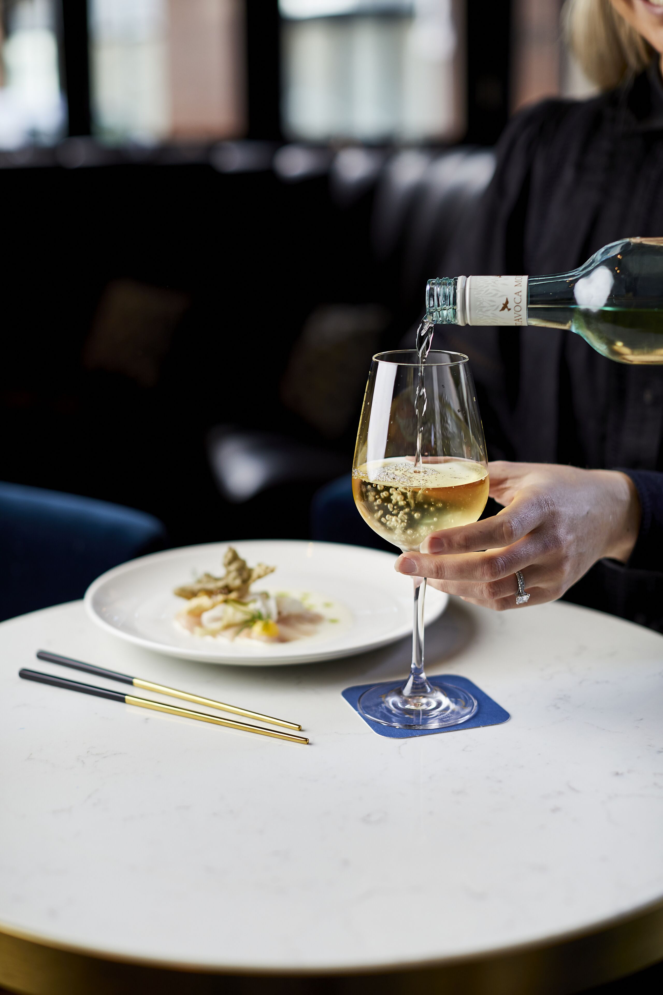 A person pouring wine into a glass on a table with a plate of food and chopsticks.