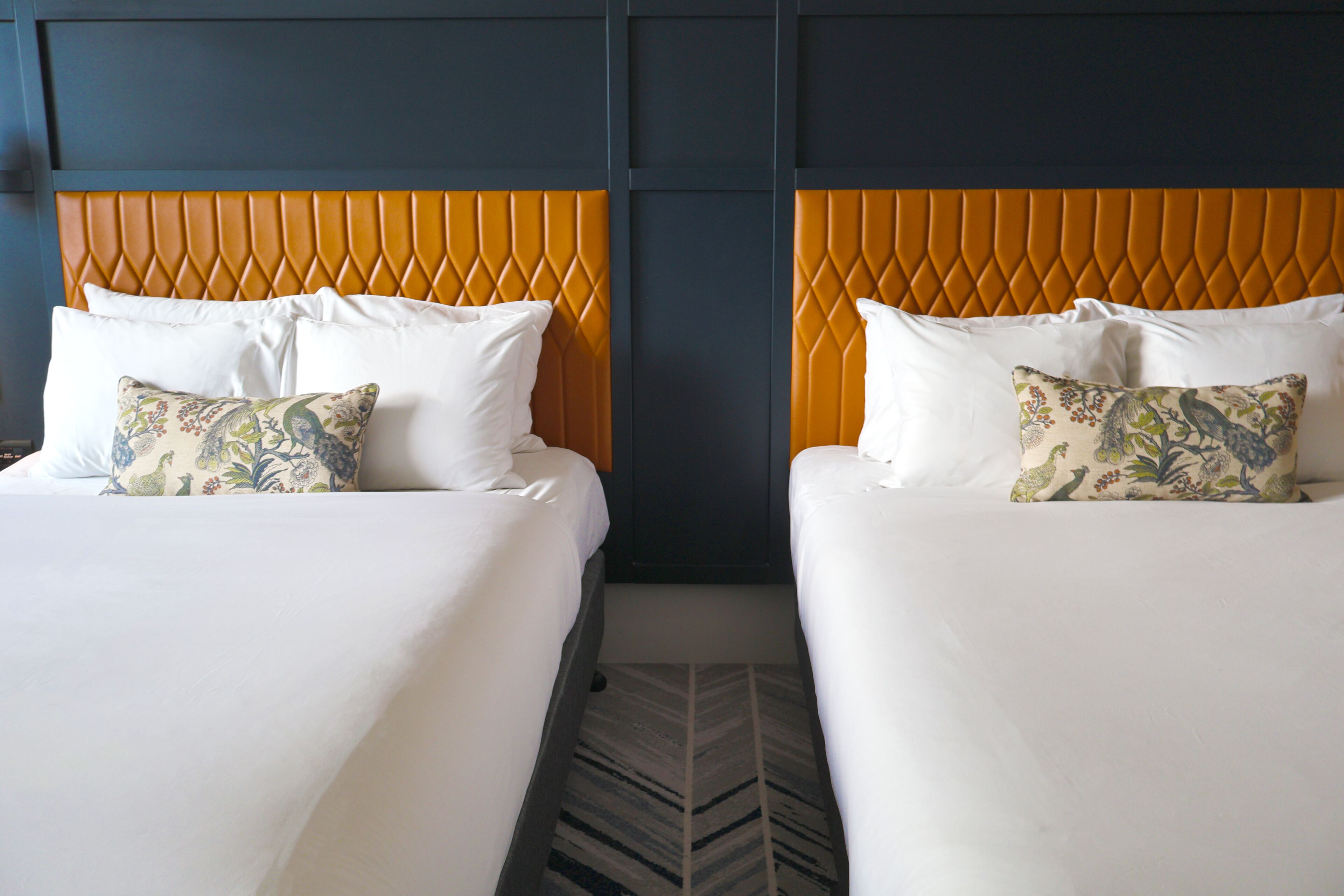 Deux lits avec du linge de lit blanc et des oreillers à motifs, un mur foncé et un tapis à chevrons.