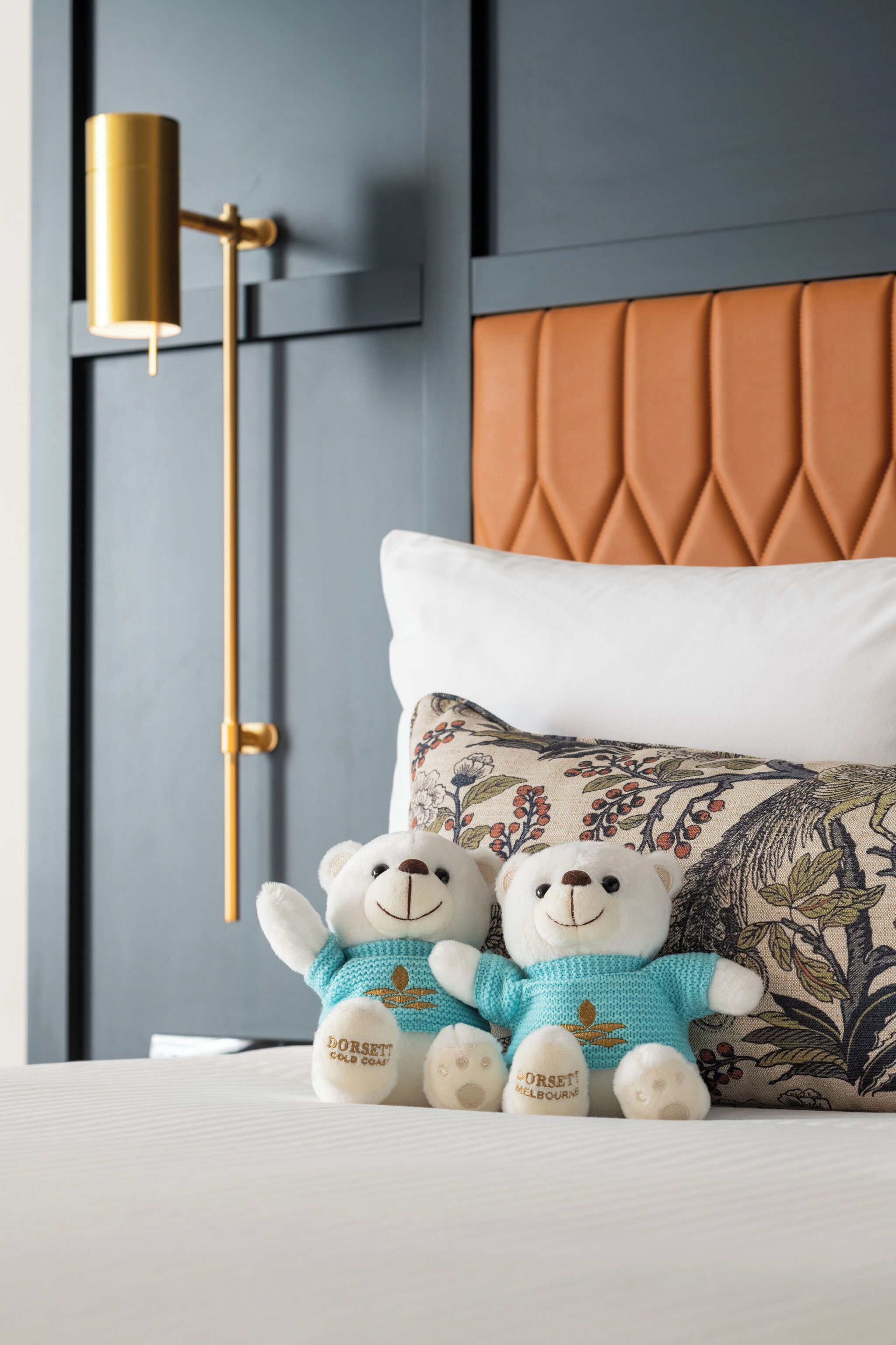 A bed with a tufted headboard, two plush teddy bears, a decorative pillow, and a modern floor lamp.
