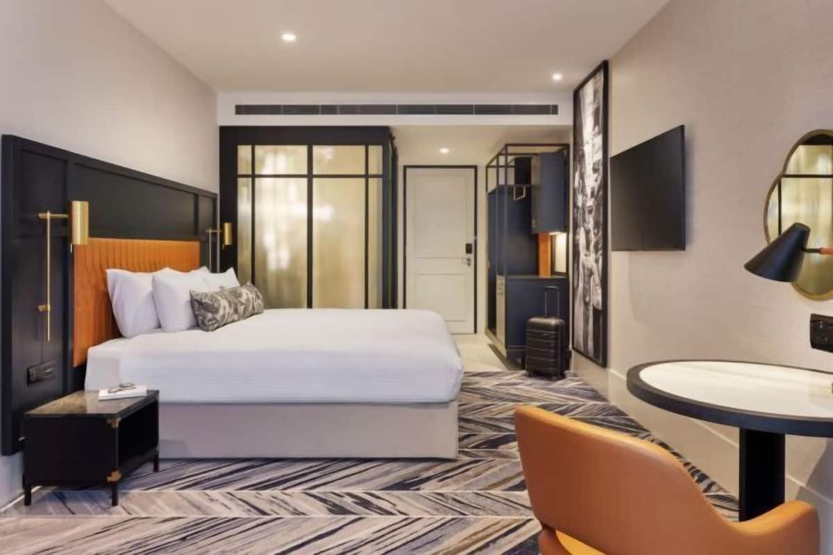 A hotel room with a bed, a bedside table, a chair, a round table, a mirror, and a wall-mounted TV.