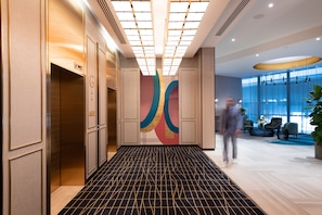 A modern hotel lobby with a geometric patterned floor, a large abstract wall art piece, and a seating area with a large window providing a view of the outdoors.