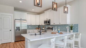 A modern kitchen with white cabinets, stainless steel appliances, a large island with bar stools, and pendant lighting. - Array of Sunshine Beach House (St. Augustine)