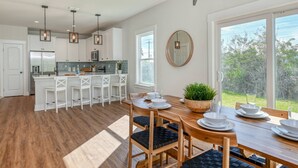 A modern kitchen with white cabinets, a central island with bar stools, and a dining area with a wooden table and chairs. - Array of Sunshine Beach House (St. Augustine)