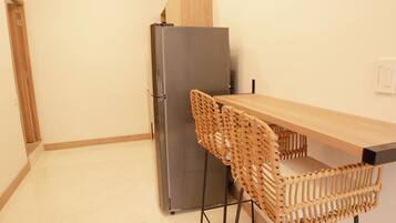 A compact kitchen with a stainless steel refrigerator, a wooden table, and a woven chair.