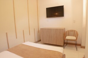 A bedroom with a bed, a TV mounted on the wall, a wooden wardrobe, and a wicker chair.