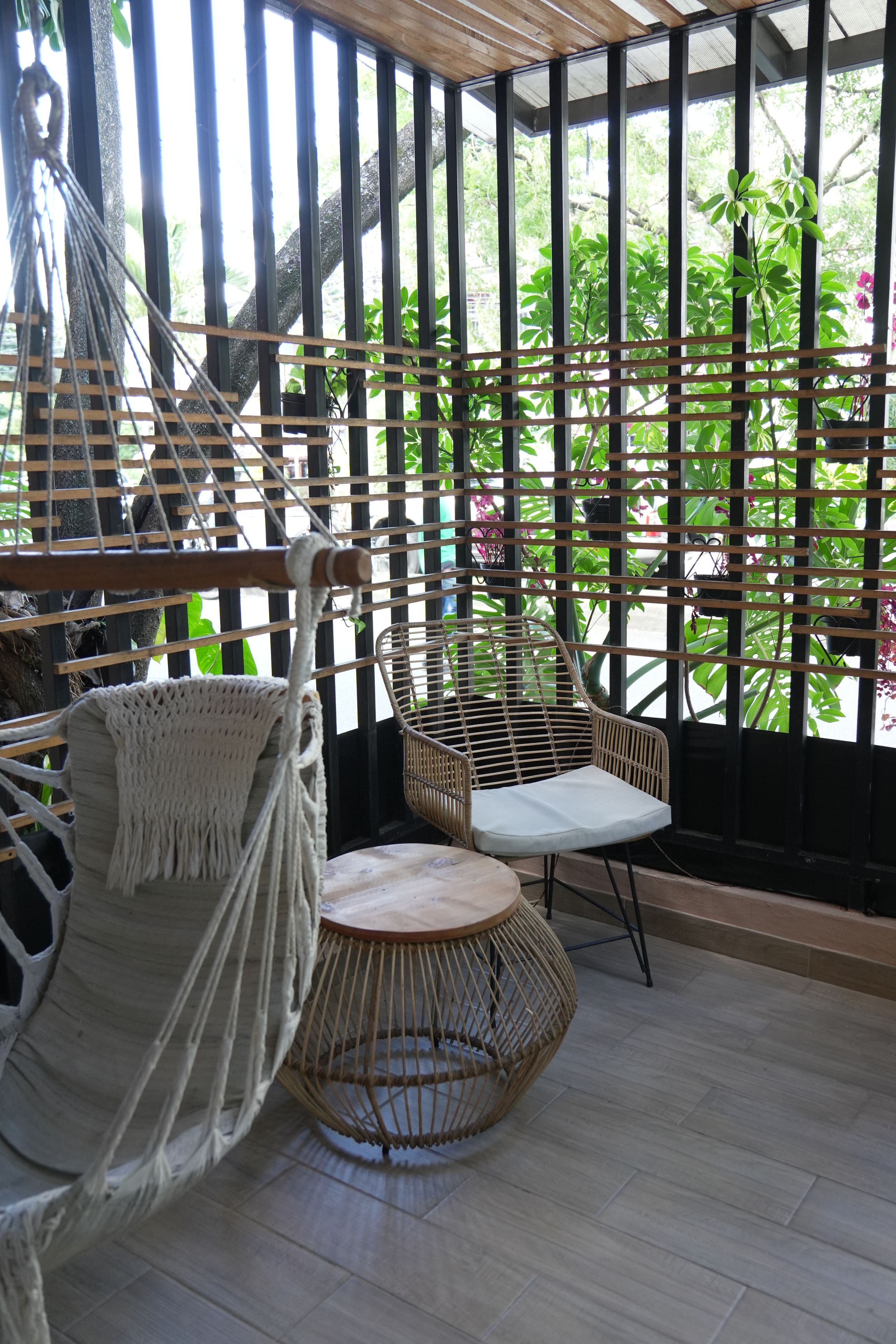 A hammock chair and a wicker chair in a room with a view of greenery through a lattice window.