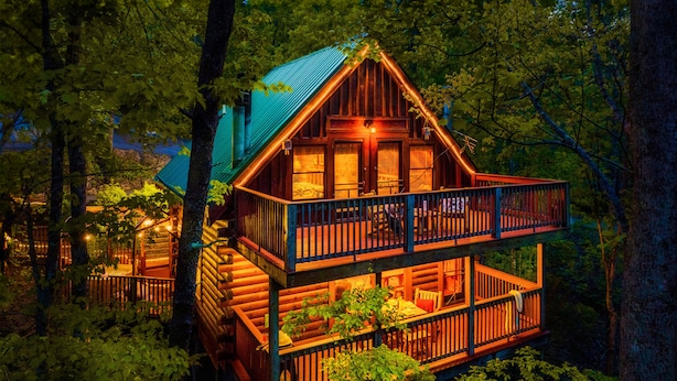 A two-story log cabin with a balcony, surrounded by dense forest. - Cabin in a gated resort with HOT TUB, RESORT POOL, BBQ GRILL, WOOD FIREPLACE  (Pigeon Forge)