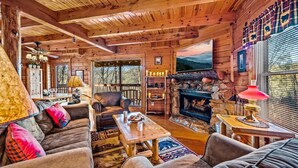 A cozy cabin interior with a fireplace, wooden furniture, and a scenic view through the windows. - Cabin in a gated resort with HOT TUB, RESORT POOL, BBQ GRILL, WOOD FIREPLACE  (Pigeon Forge)