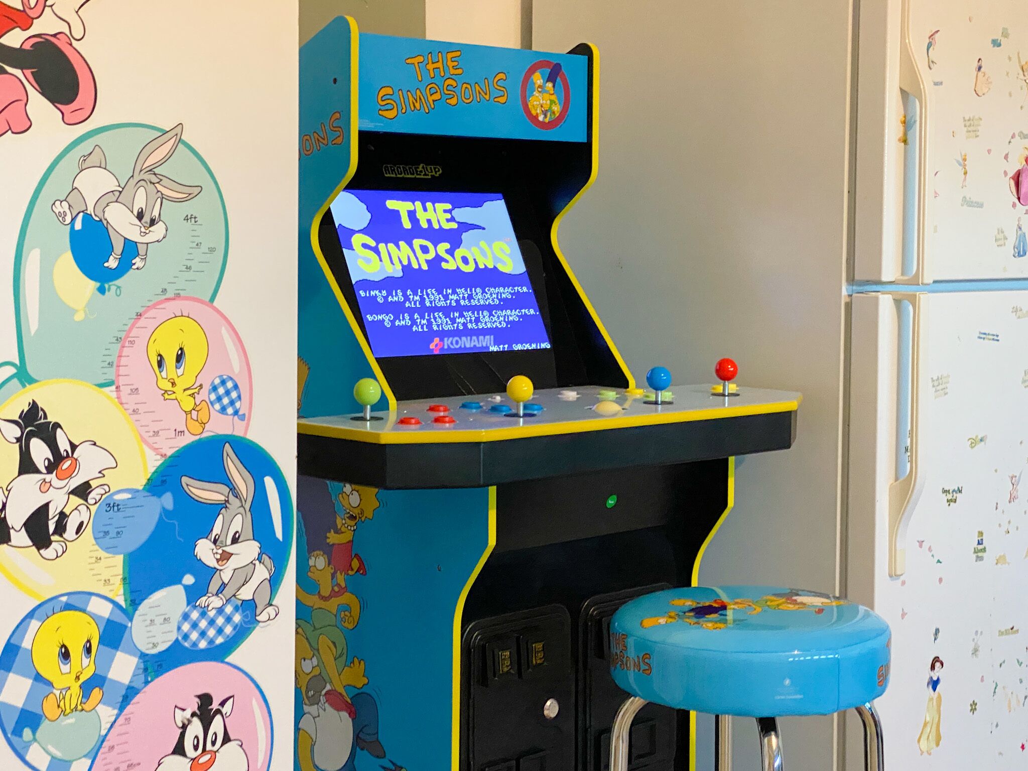 A The Simpsons arcade game with a blue stool and cartoon stickers on the wall.
