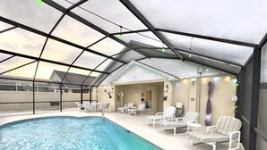 An indoor swimming pool with clear water, surrounded by white lounge chairs and a covered patio area with string lights.