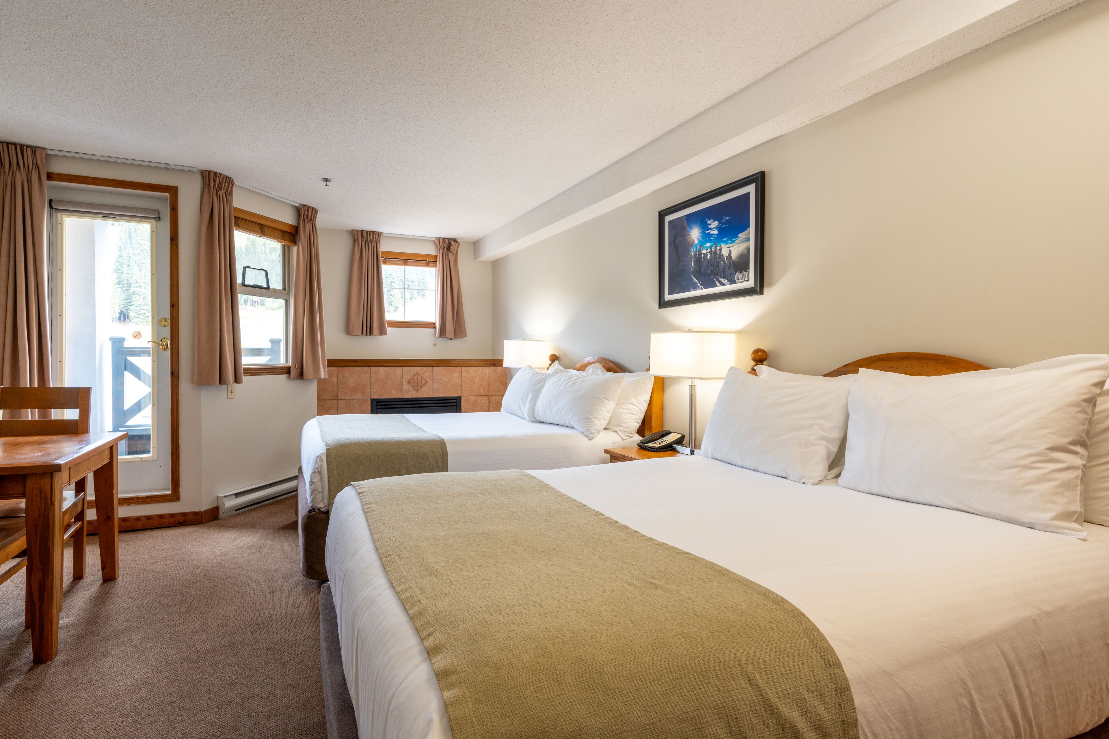 A hotel room with two beds, a desk, a chair, a window with curtains, a framed picture on the wall, and a lamp on the nightstand.