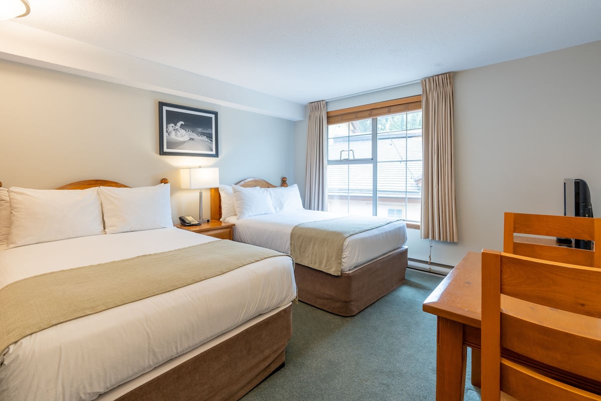 Ett hotellrum med två sängar, ett skrivbord, en stol, ett nattduksbord, en lampa, en tavla på väggen och ett fönster med gardiner.