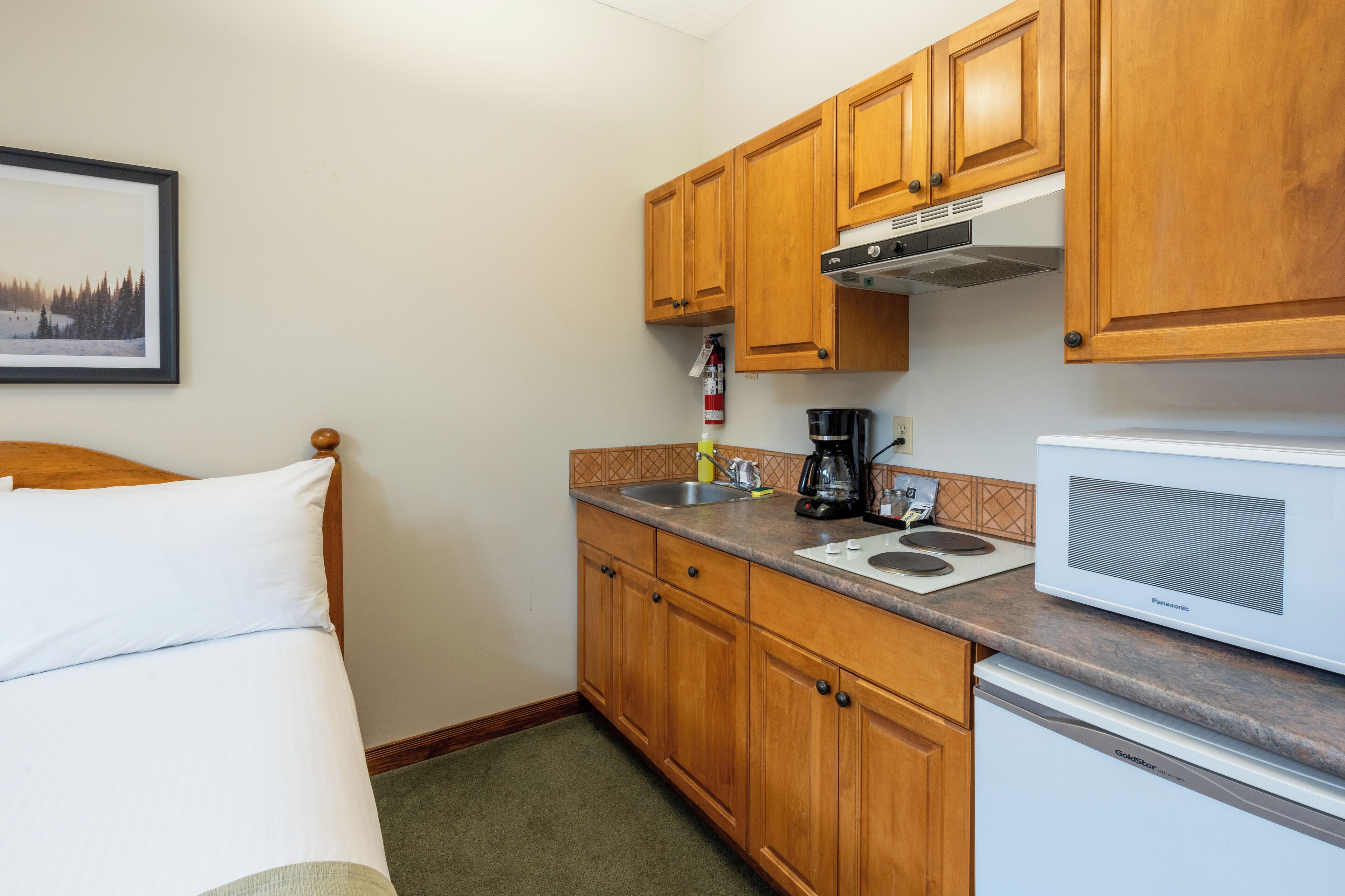 Habitación de hotel pequeña con armarios de madera, cama, microondas, cafetera y lavamanos.