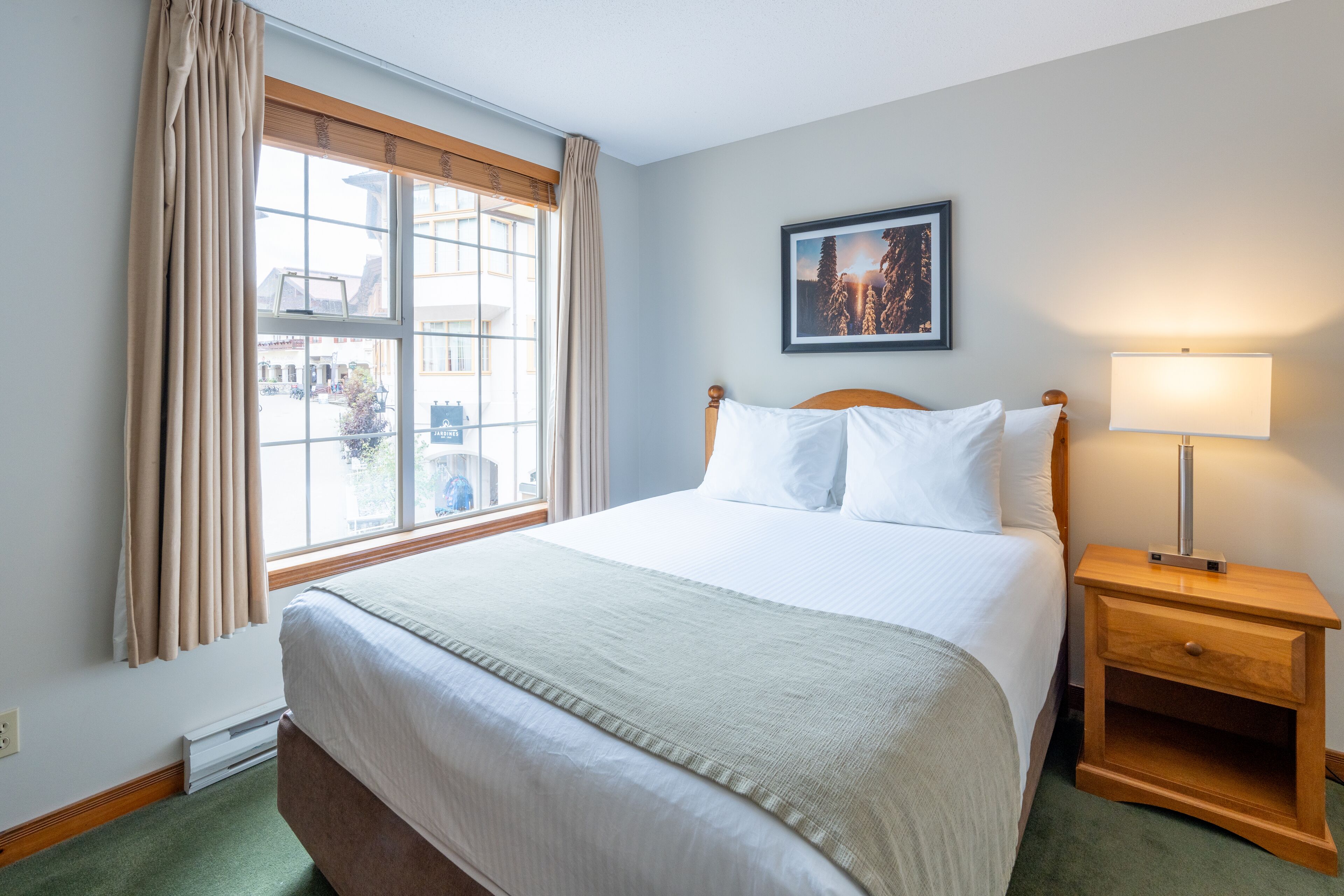 Una habitación de hotel con una cama grande, una mesita de noche con lámpara, una ventana con cortinas y un cuadro enmarcado en la pared.