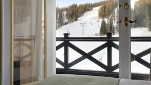 Une chambre avec vue sur une pente de montagne enneigée, un lit et une fenêtre ornée d’une rambarde en métal.