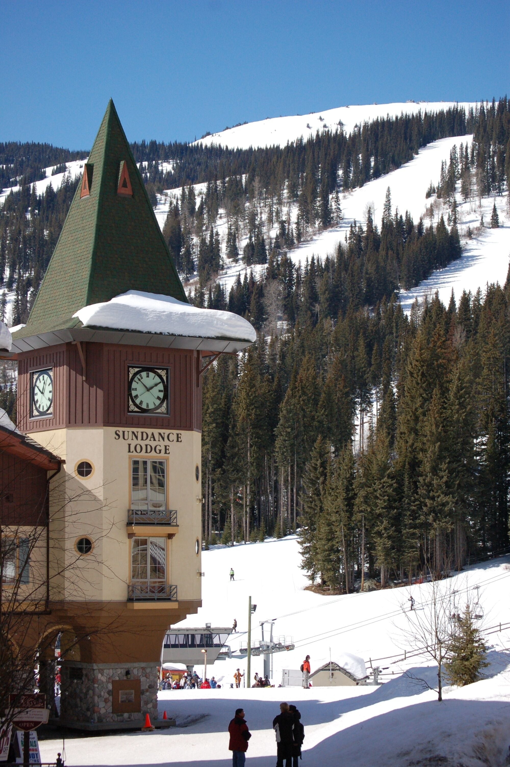 A lodge with a clock tower, surrounded by snow-covered mountains and trees.