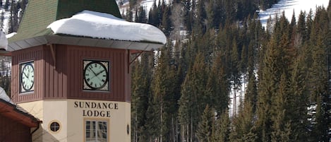 A lodge with a clock tower, surrounded by snow-covered mountains and trees.