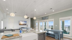Une cuisine dotée d’un grand îlot central, un coin repas offrant une vue agréable et une enseigne sur laquelle on peut lire « BEACHHOUSE ».