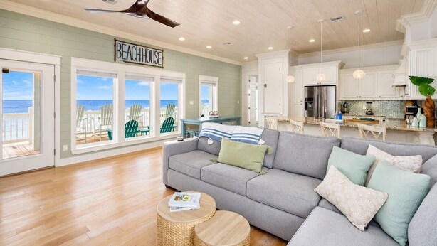 Un salon spacieux avec un canapé gris, un ventilateur de plafond et une vue sur la plage grâce à de grandes fenêtres.