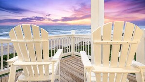 Deux chaises Adirondack blanches sur une terrasse en bois, avec vue sur l’océan et un coucher de soleil.