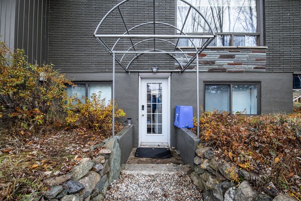 Ein modernes Gebäude mit Steineingang, einer Metallpergola über der Tür und einem blauen Gegenstand auf der rechten Seite.