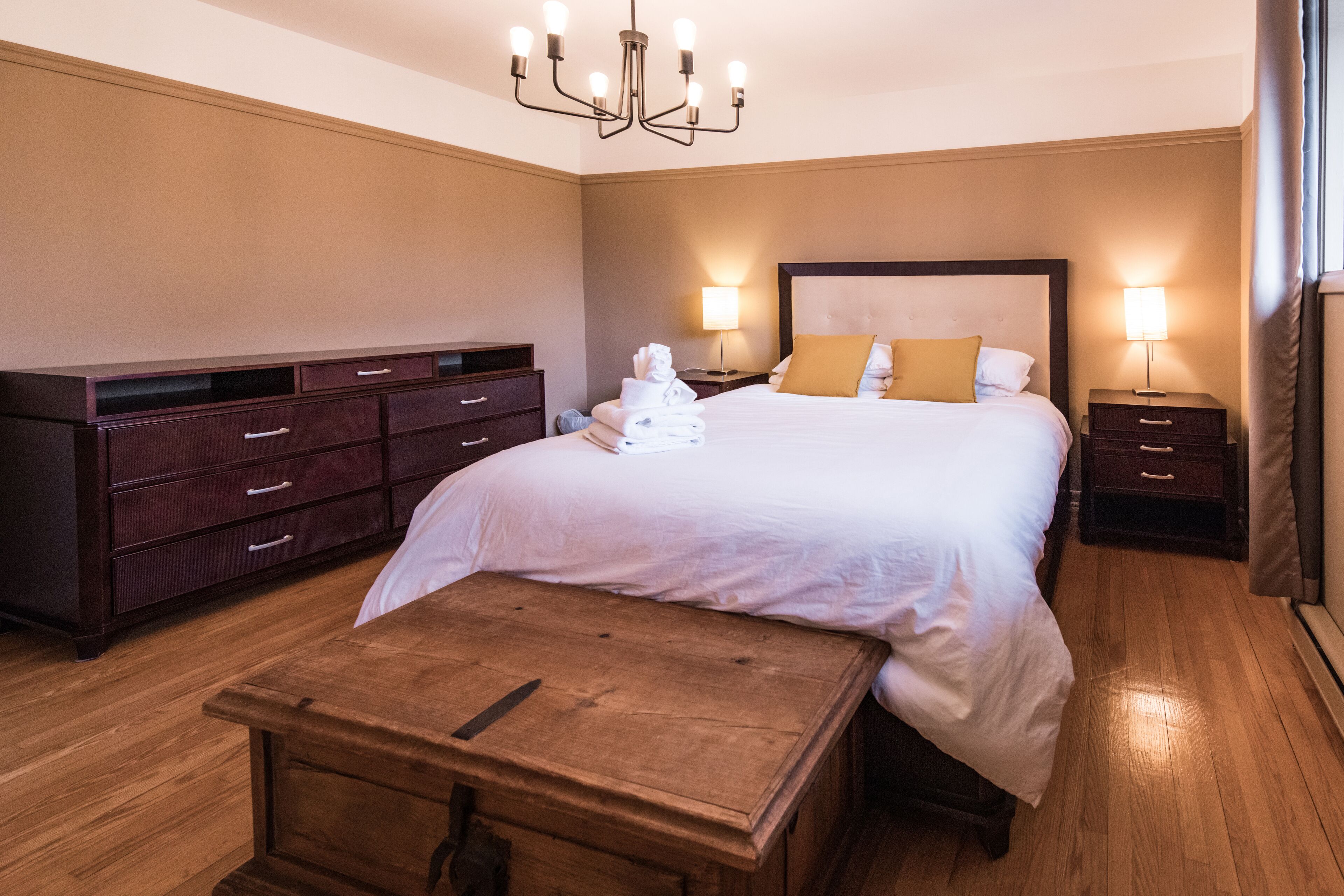 A hotel room with a large bed, wooden nightstands, a wooden bench, and a chandelier.