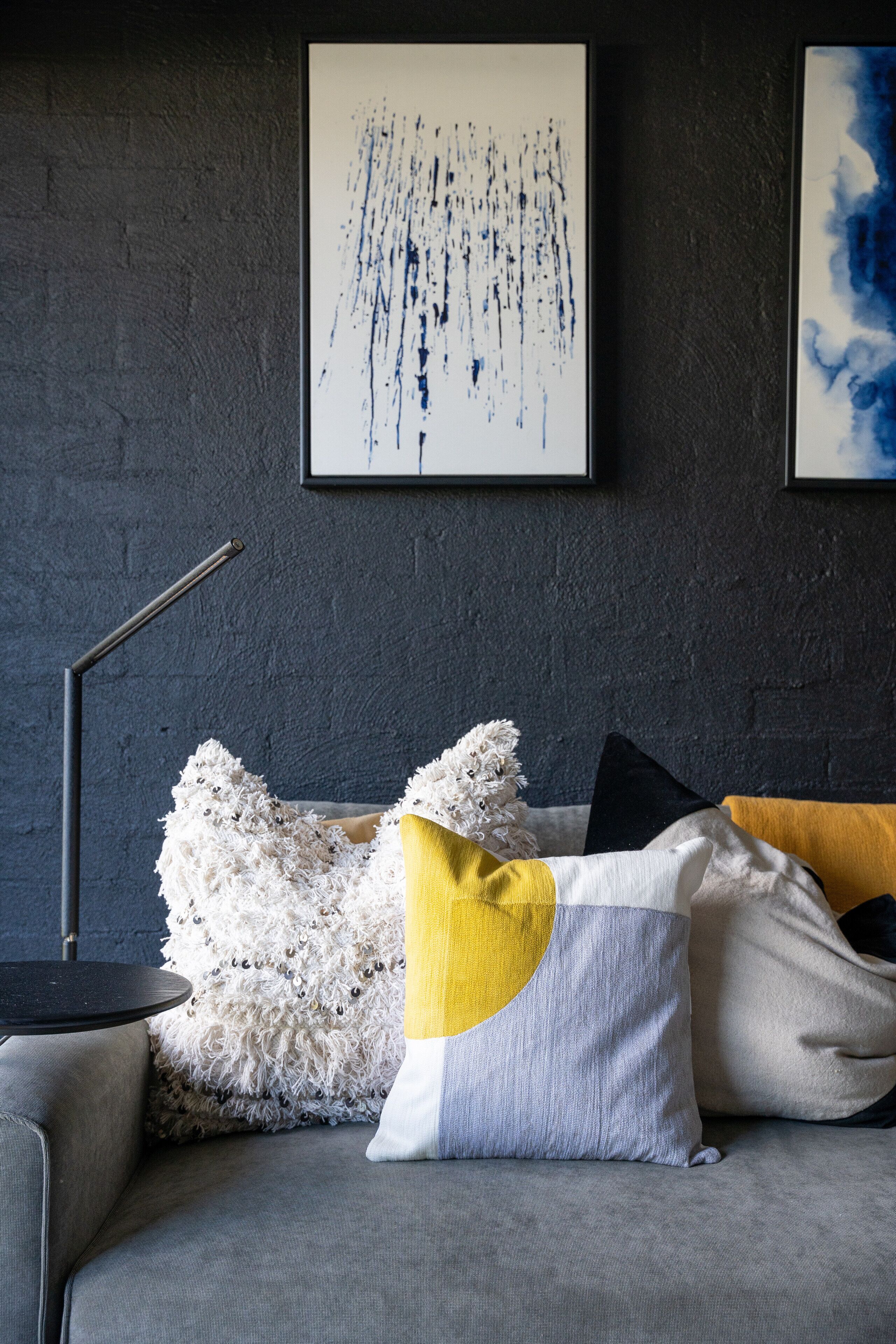 A modern living room with a grey sofa, a fluffy white pillow, and a yellow and white pillow. There are two framed abstract paintings on the wall.