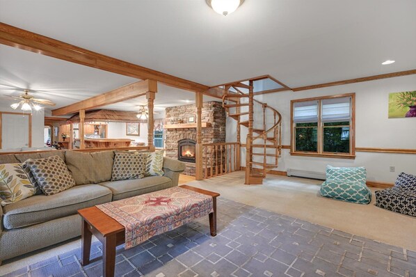 A cozy living room with a stone fireplace, wooden beams, a spiral staircase, and a large window. - Muhlenberg Cottage | Mount Gretna (Mount Gretna)