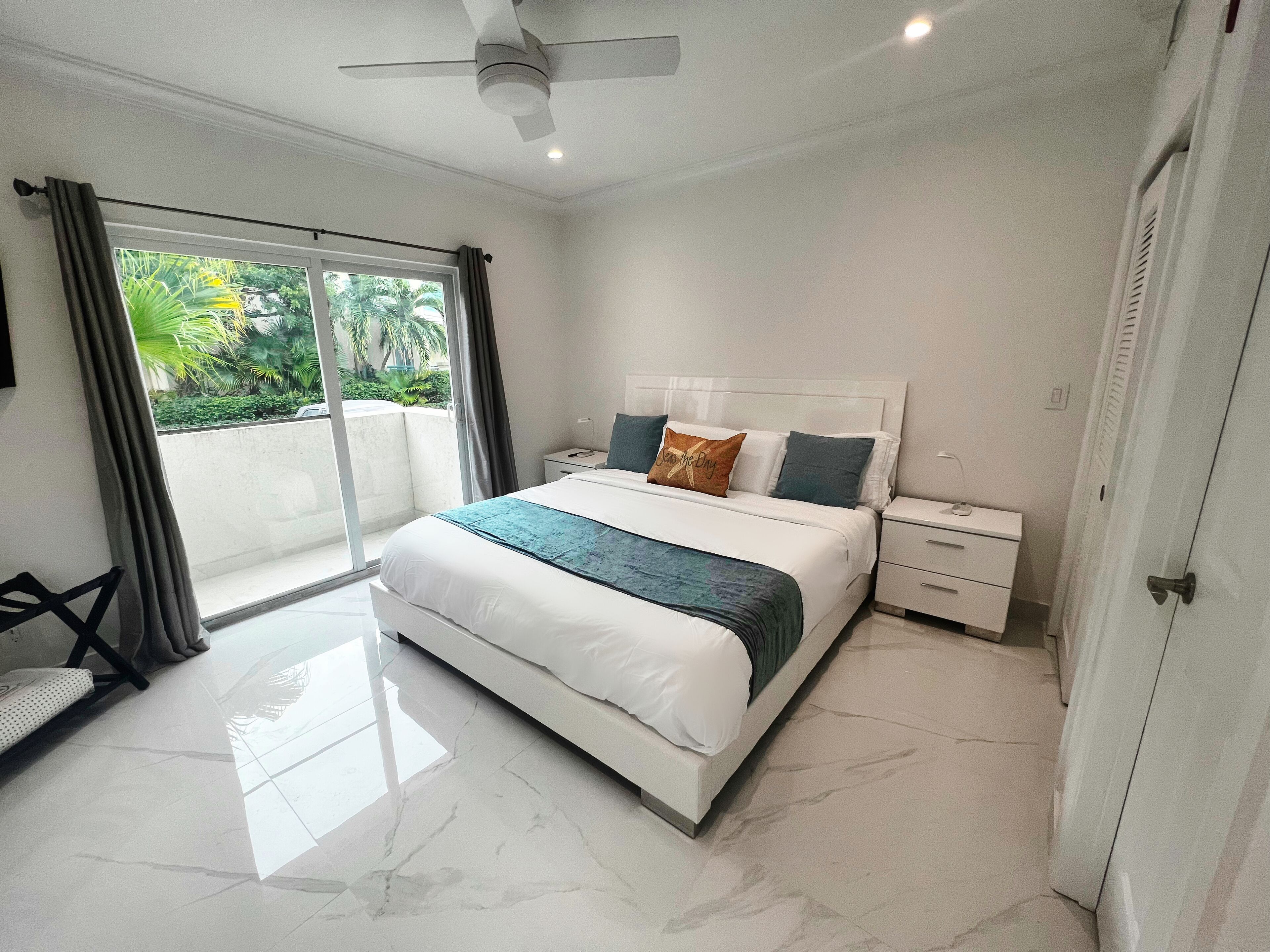 Une chambre moderne avec un grand lit, une table de chevet, un ventilateur de plafond et une vue sur la verdure environnante.