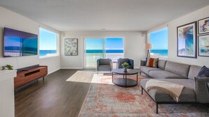 A modern living room with a grey sofa, a coffee table, and a TV mounted on the wall. There is a large window with a view of the ocean.