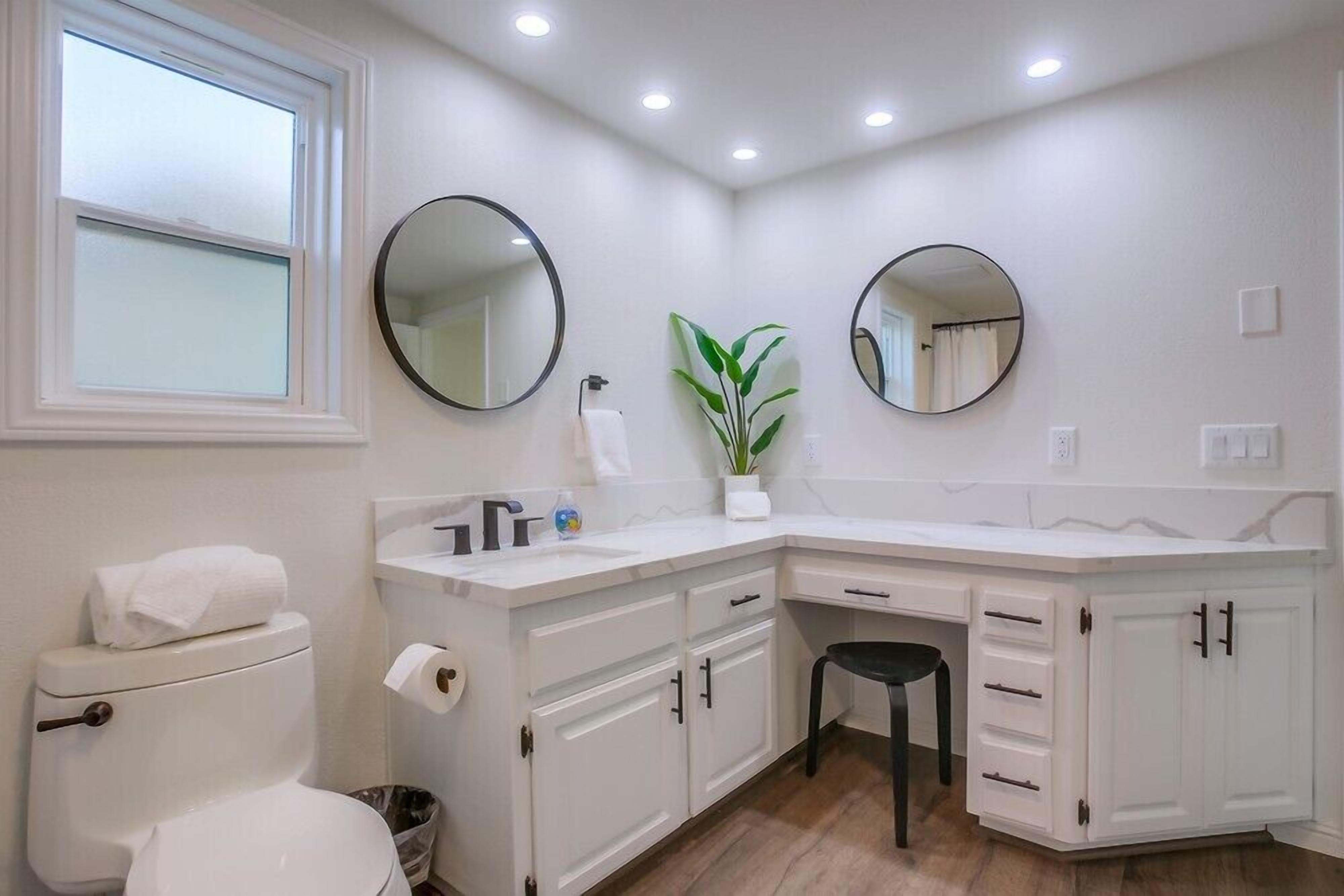A bathroom with white cabinetry, two round mirrors, a sink, a toilet, and a potted plant.