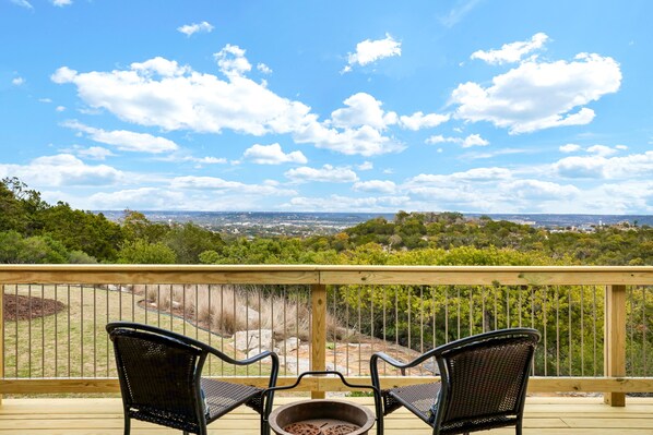 Une terrasse en bois avec deux fauteuils en osier noir, placés face à un foyer extérieur, et offrant une vue panoramique sur un paysage verdoyant, avec des arbres et des bâtiments situés au loin.
