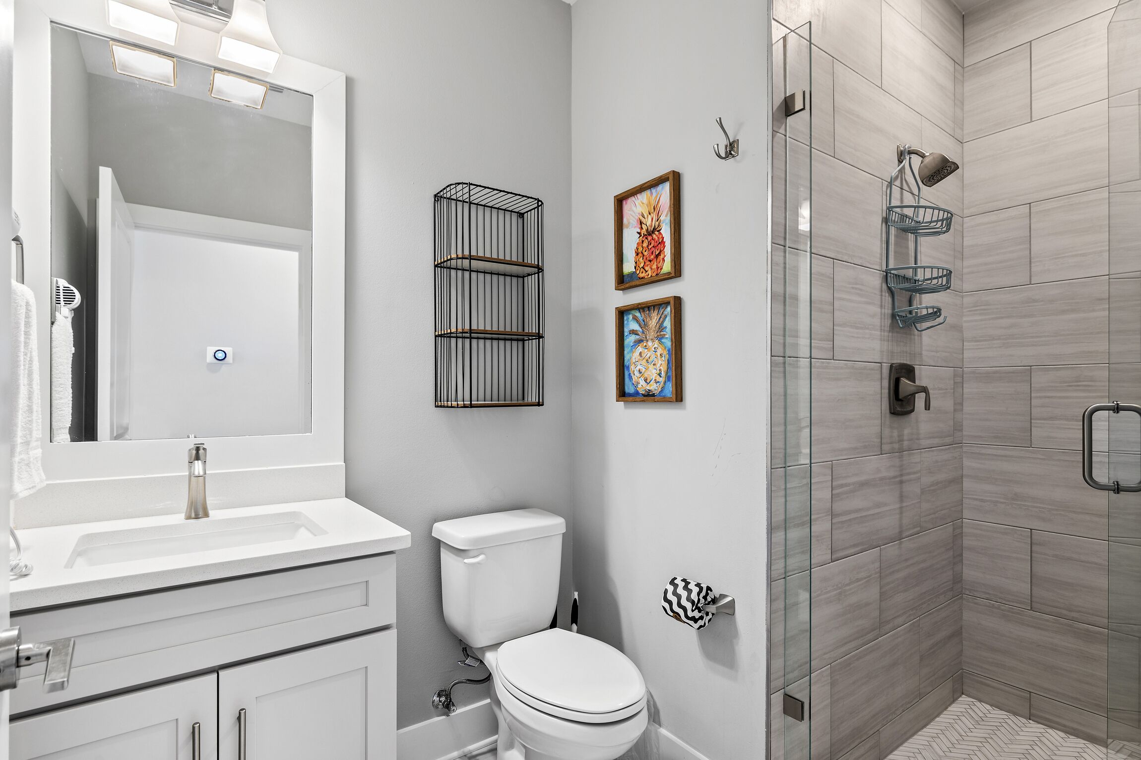 Une salle de bain moderne avec un lavabo, des toilettes et une douche, le tout de couleur blanche. Elle comprend un miroir, un porte-serviettes et des œuvres d’art encadrées accrochées au mur.