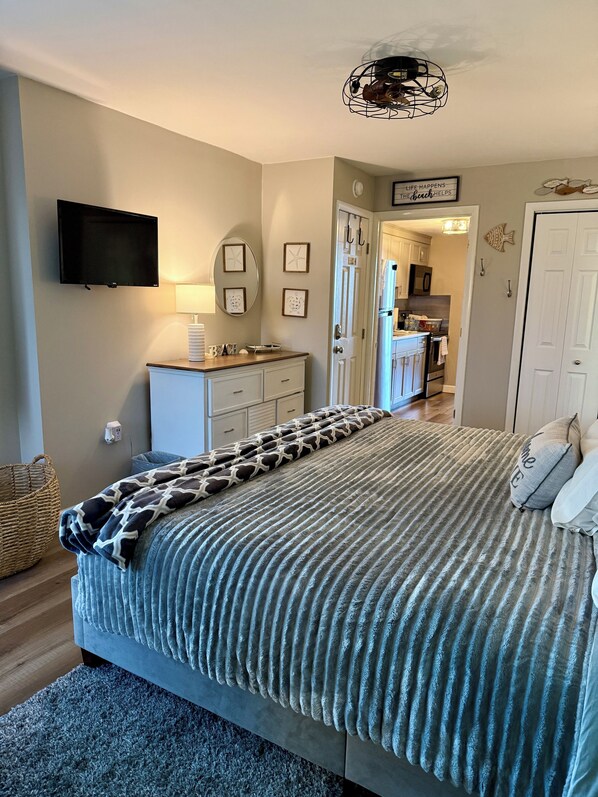 A bedroom with a bed, a dresser, a television, and a kitchen visible through an open door.
