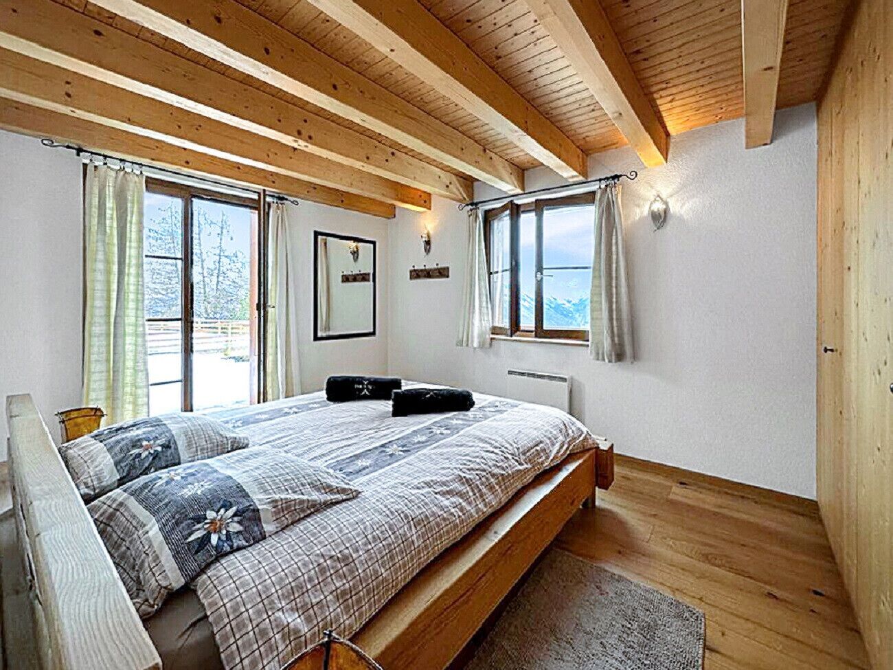 Un dormitorio con techo de madera, una cama grande con colcha estampada, dos lámparas de pared y un ventanal con cortinas.