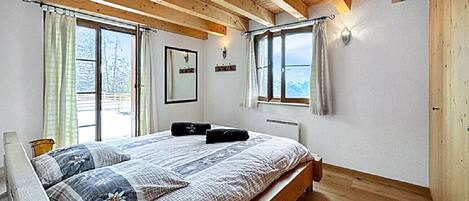 Un dormitorio con techo de madera, una cama grande con colcha estampada, dos lámparas de pared y un ventanal con cortinas.