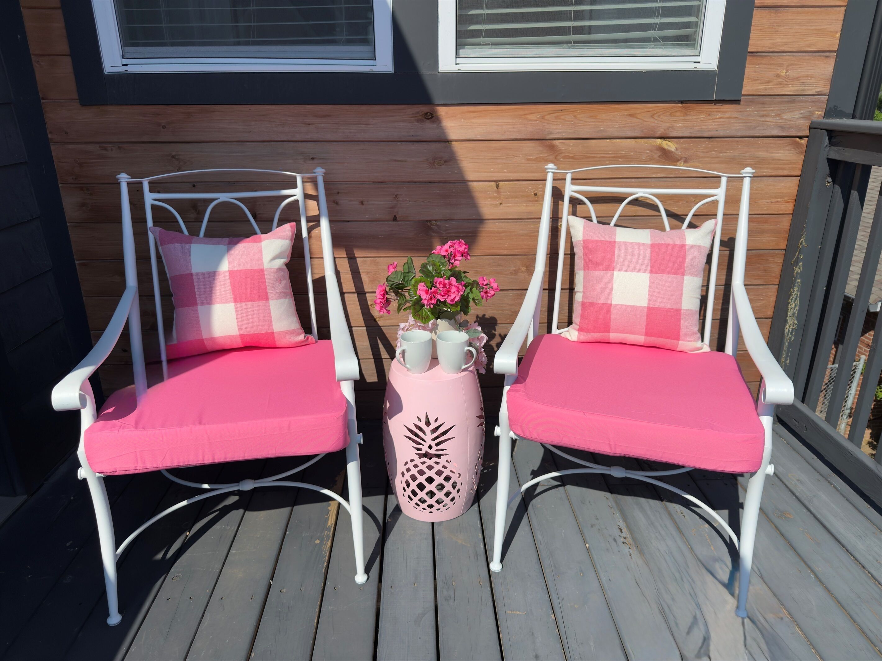 Deux chaises en métal blanc, dotées de coussins roses et d’oreillers à carreaux, sont placées sur une terrasse en bois.