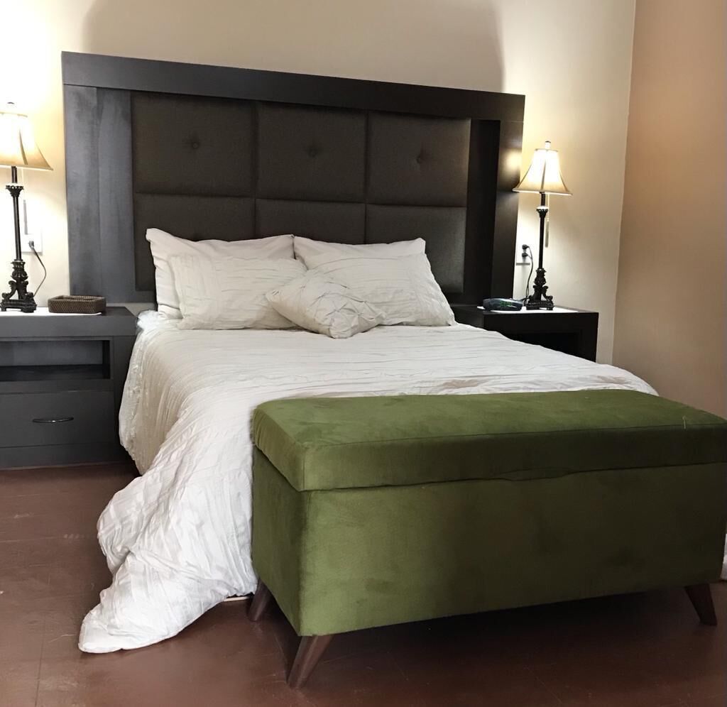 Una cama bien tendida con ropa blanca y un banco tapizado en verde, un cabecero oscuro y dos mesitas de noche con lámparas.