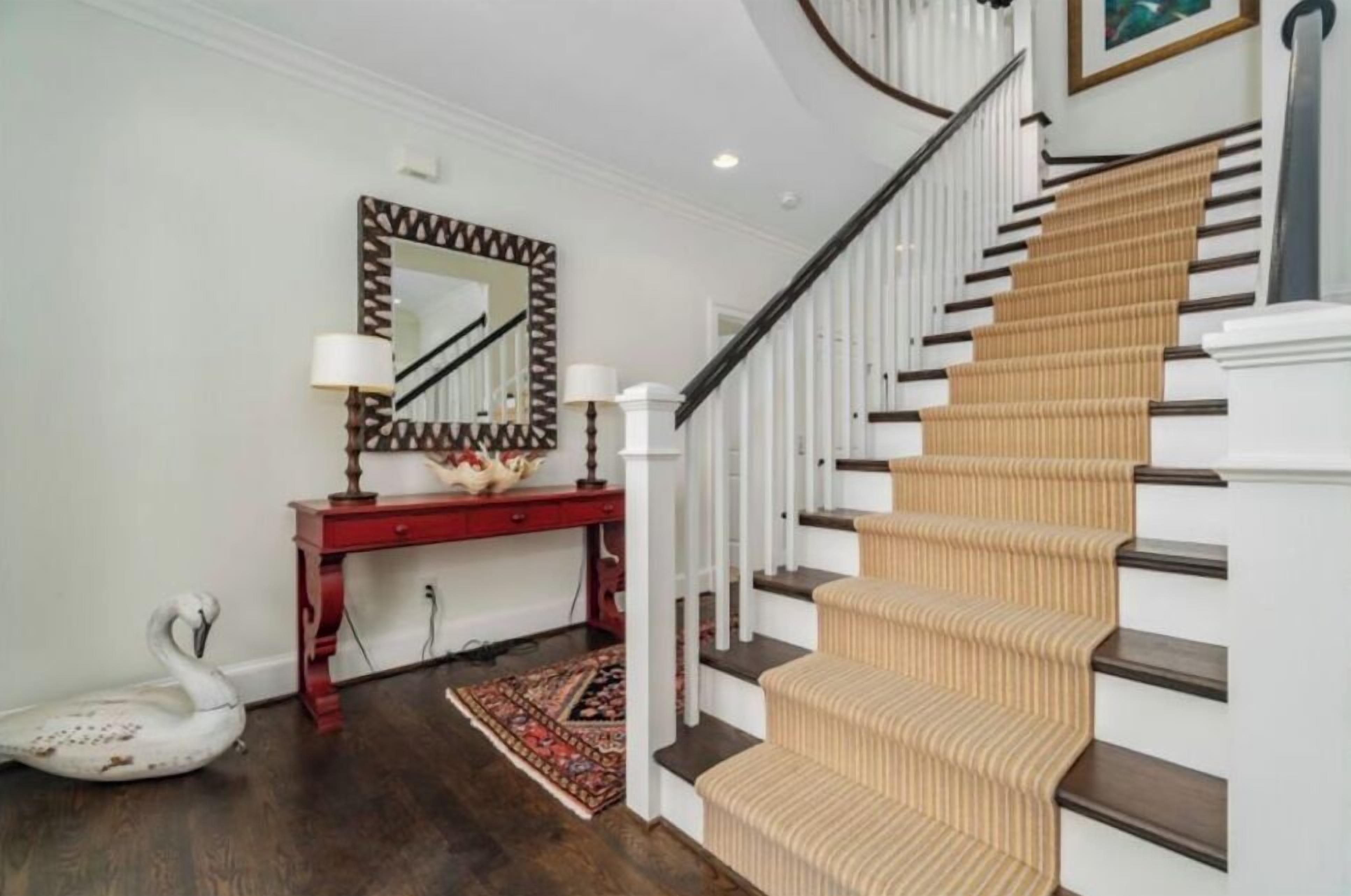 Un escalier recouvert d’une moquette beige, avec des marches en bois foncé, menant à un palier où se trouve une console rouge et un miroir à cadre géométrique.