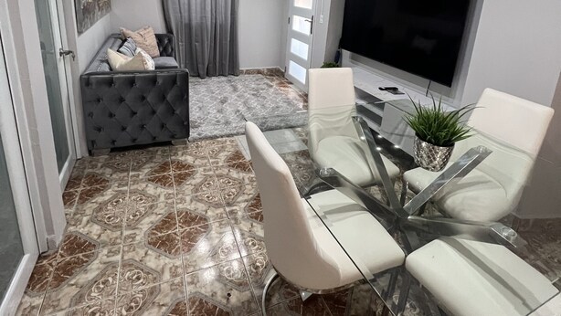 Ein modernes Wohnzimmer mit einem grauen Sofa, einem Glastisch mit Stühlen, einem Flachbildfernseher und einem Bodenbelag mit Muster.