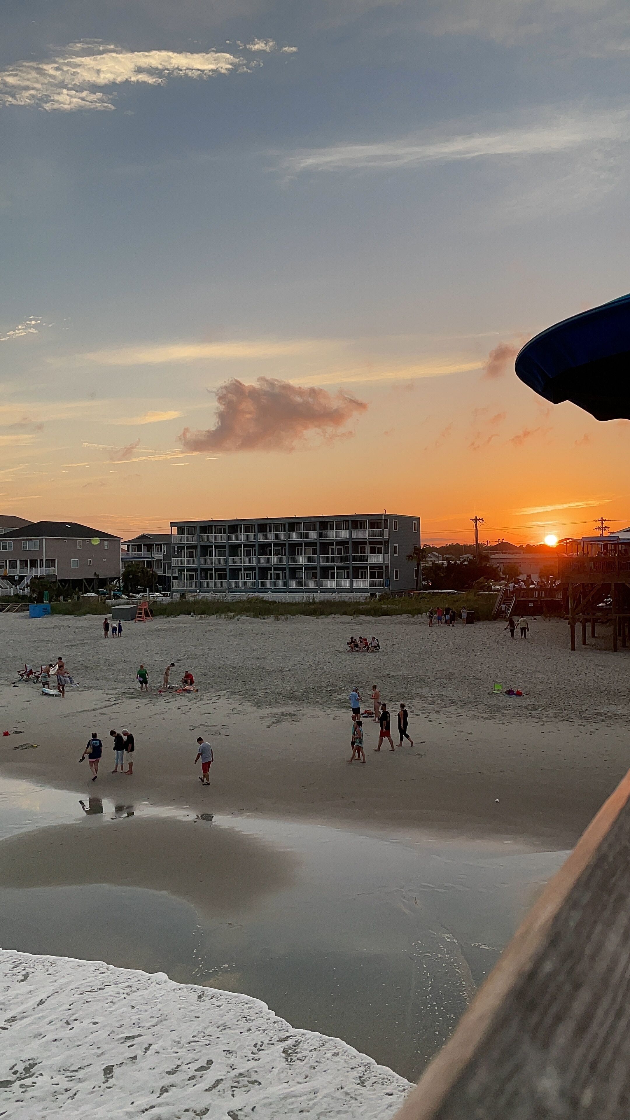 Ein Strand mit Menschen, die laufen und stehen, Gebäuden im Hintergrund und einem Sonnenuntergang.