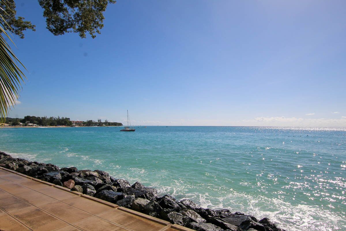 A coastal view with a rocky shoreline, clear blue water, and a sailboat in the distance.